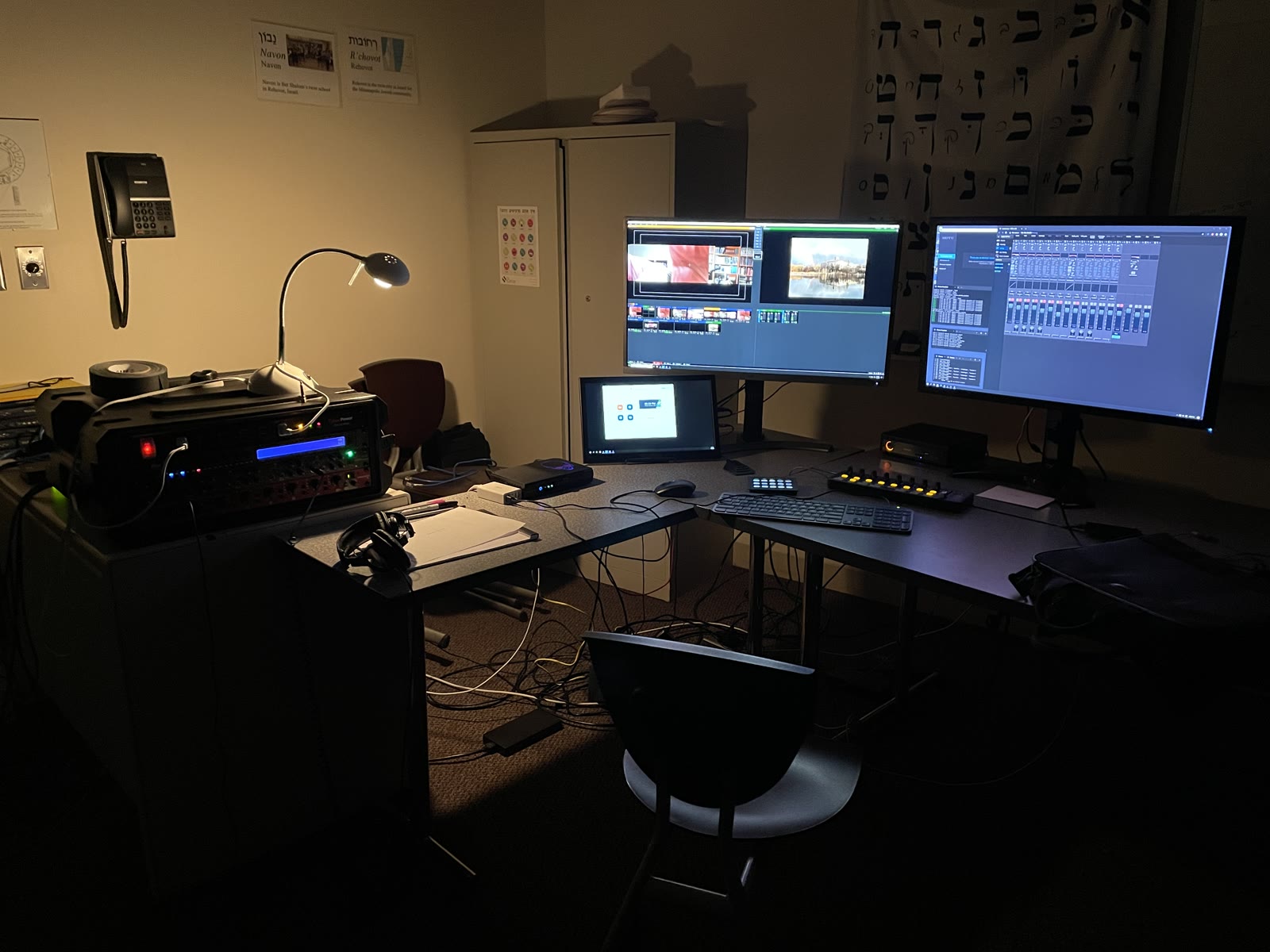 A synagogue office turned broadcast control room at night: a vMix multiview and an audio routing grid glowing on two monitors, a Stream Deck and rack mixer lit by a desk lamp, headphones on a legal pad, an empty chair, a Hebrew alphabet chart on the wall