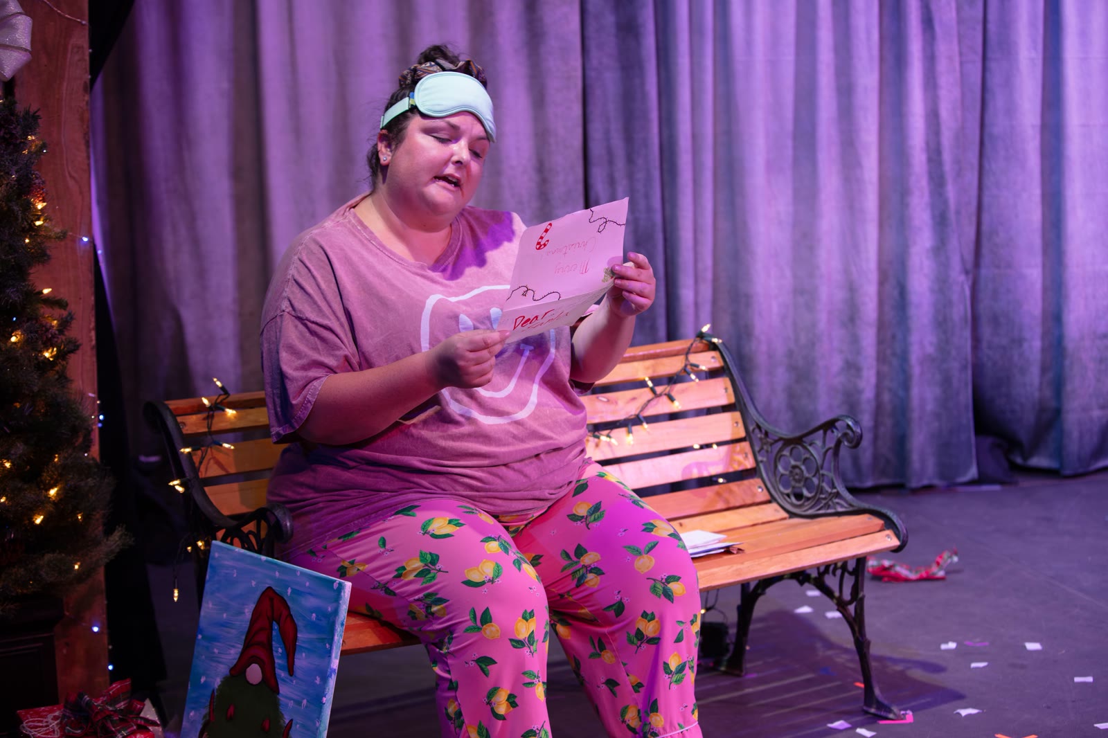 A woman in pyjamas and a sleep mask sits on a park bench reading a card; fairy lights are draped along the bench and a lit tree stands at the edge of frame, confetti snow across the floor