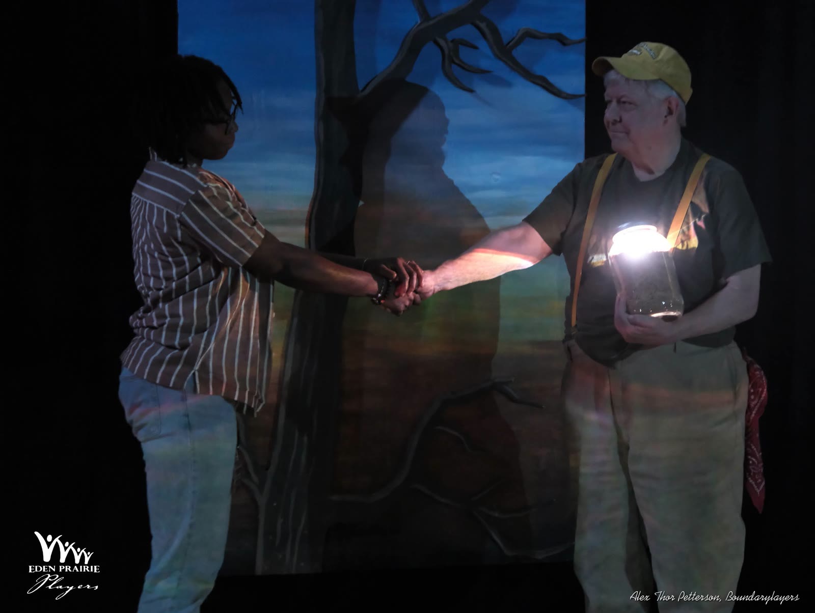 Two actors clasp hands in low blue light against a projected tree at dusk; one holds a jar glowing like a held flame