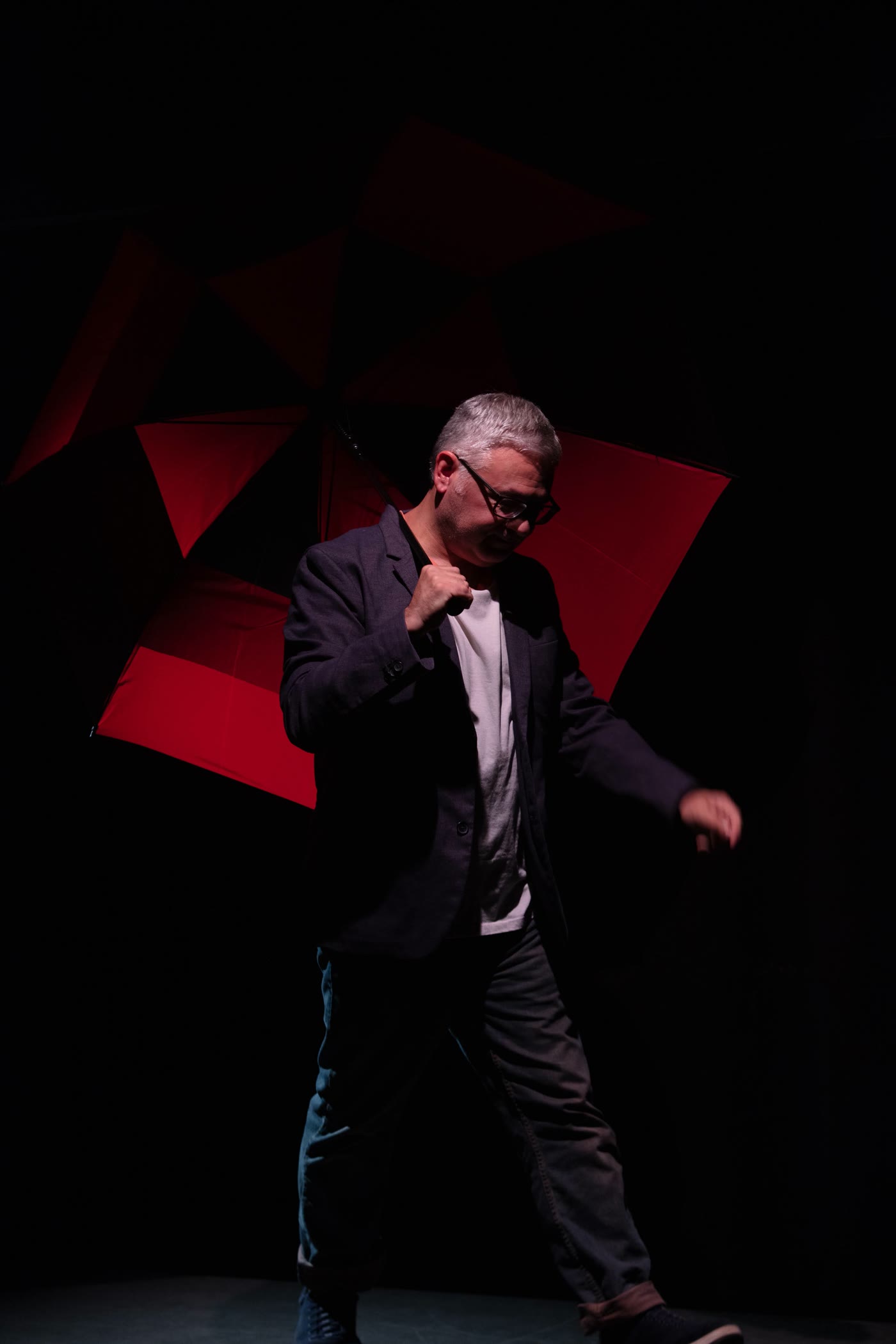 A man in a dark jacket walks through a tight pool of light; behind him a large red-and-black umbrella glows out of total darkness