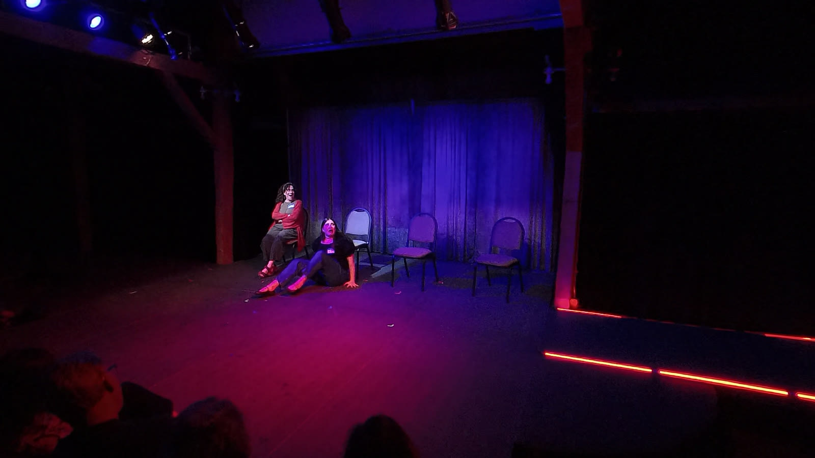 A dark stage washed blue behind a hanging curtain: one performer sits in a chair, another on the floor looking upward, four empty chairs beside them — and two lines of red-orange light run along the downstage edge, out of frame into the dark