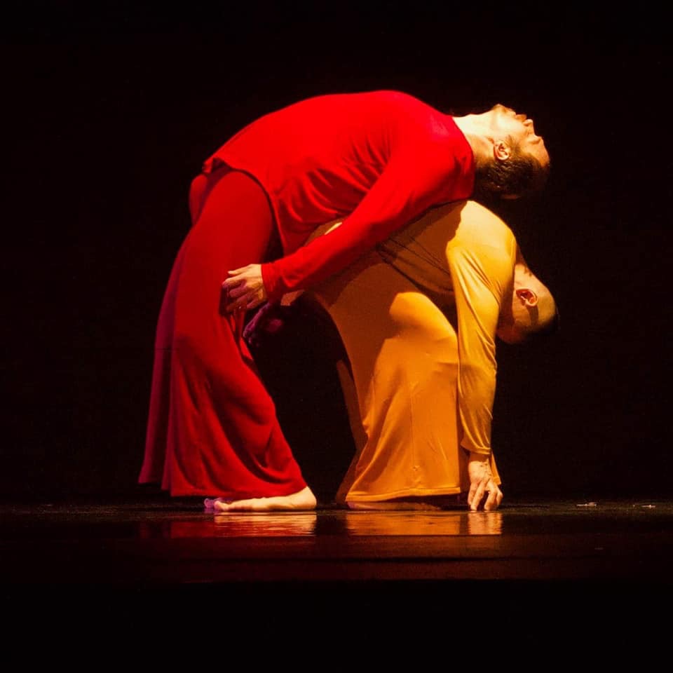 Two dancers against pure black: one in deep red arches backward over one in gold, both lit so the colour is the only thing in the frame