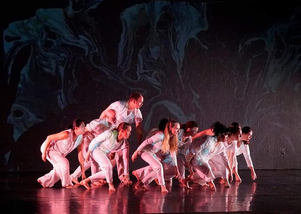 Ten dancers in white crouched along a diagonal, mid-crawl, with a vast marbled painting projected across the back wall and spilling onto them