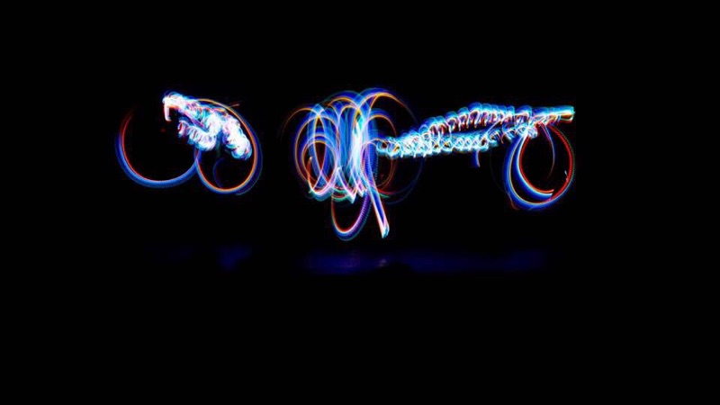 A long exposure on a black stage: loops and helices of blue, white and red light drawn in the air by moving sources, with no dancer visible — only the path each one travelled