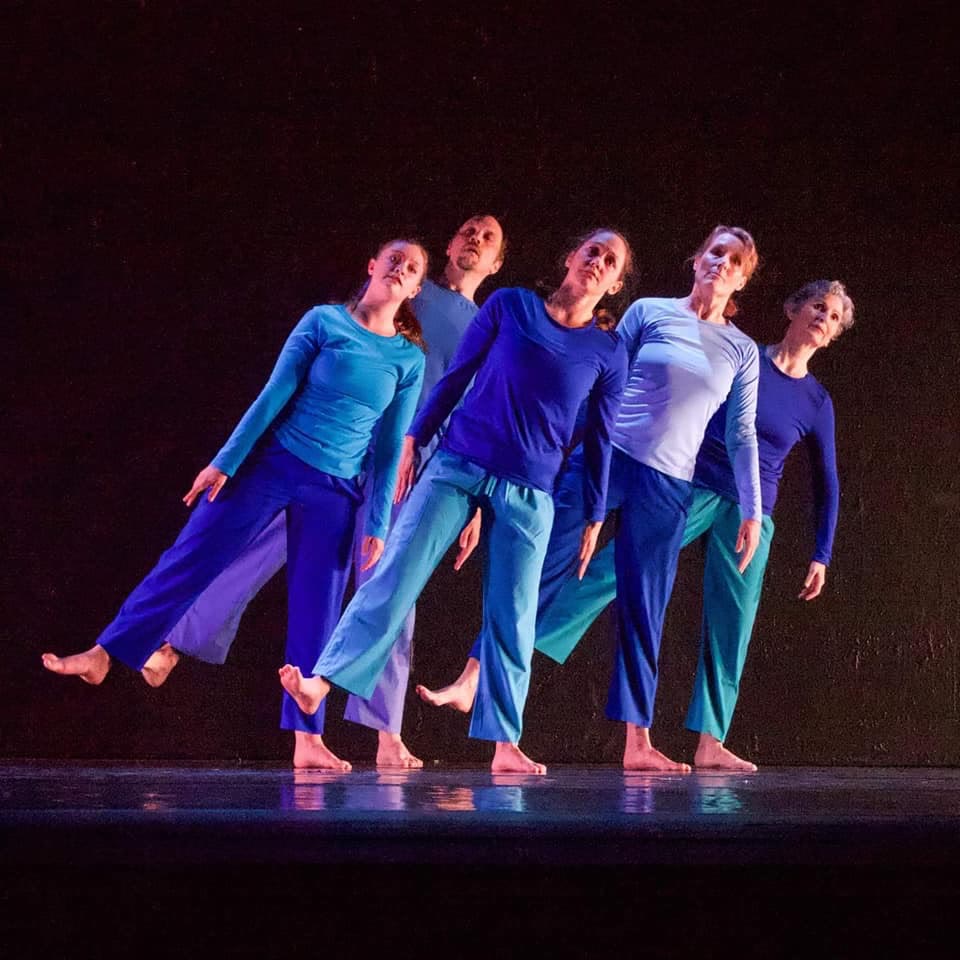Five dancers in blues, staggered and leaning together, their reflections held in the floor
