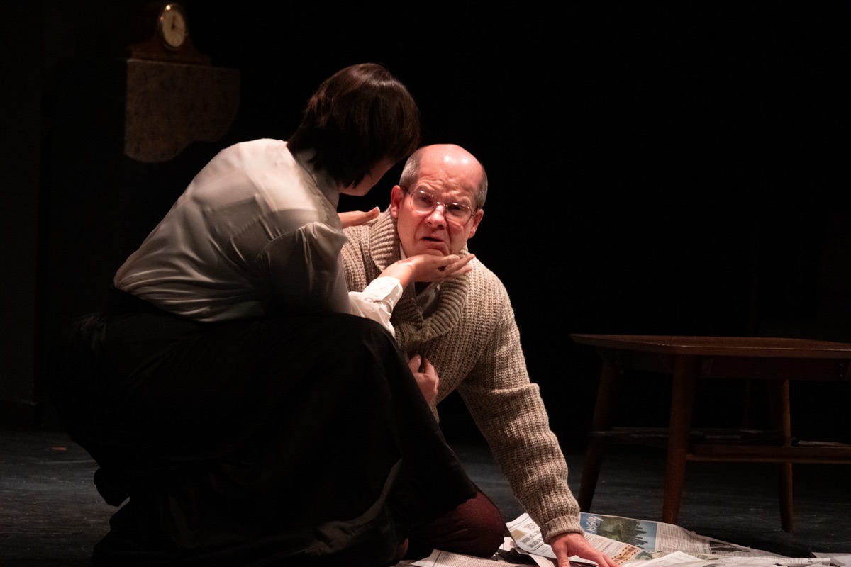 In tight light, a kneeling figure cups the chin of a frightened man on the floor among scattered newspapers